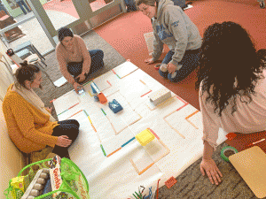A group of teachers from New Bedford Public Schools are sitting on the floor around a large white paper. They have used popsicle sticks, stick notes, and small recyclables to design a city scape for the Bee-Bot robot to nagivate. They are engaged and laughing, working together.